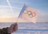 A hand holding an Olympic flag against a snowy landscape during sunset