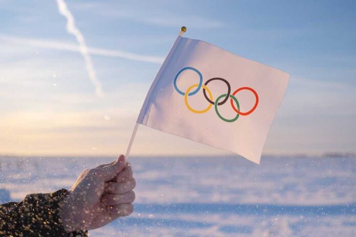 shutterstock_2097149272.jpg A hand holding an Olympic flag against a snowy landscape during sunset