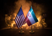 Flags of the United States and Iran displayed on stands with a smoky background
