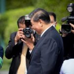 A public figure surrounded by photographers at an outdoor event