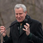 A man in a black coat speaking passionately at an outdoor event