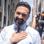 A smiling man in a light blue shirt gestures with his hand in an outdoor urban setting