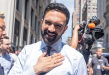 NYC Mayor WEAPONIZES City Hall Against Property Owners A smiling man in a light blue shirt gestures with his hand in an outdoor urban setting