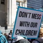 Scandal Erupts: Schools Bypass Parental Consent! Protest sign held by a crowd in an urban square