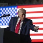 A man in a suit gestures while speaking at a podium with an American flag in the background