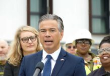 Feds Wave Through Merger; California Loads Lawsuit Man in a navy suit speaking at a microphone with people standing behind