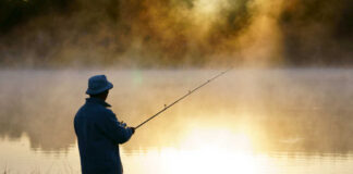 A fisherman standing by a misty lake at sunrise, holding a fishing rod