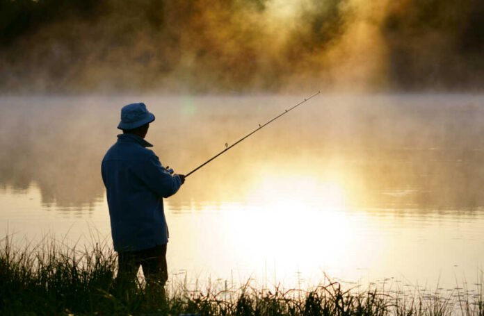 Caucasian,Fisherman,,50's,,At,The,Lake,With,Fog,,Early,Morning A fisherman standing by a misty lake at sunrise, holding a fishing rod