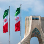 Shadow Rulers Exposed — Deals Won’t Save Iran Three Iranian flags in front of the Azadi Tower against a blue sky