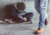 Person sitting on sidewalk with hood up beside a paper cup