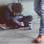 Harm Reduction or Public-Funded Paraphernalia? Person sitting on sidewalk with hood up beside a paper cup