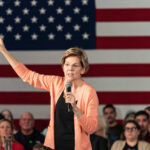 Warren’s IRS Beast: Billionaires Cornered A woman speaking into a microphone with an American flag backdrop