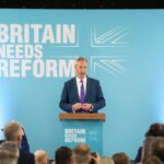 A politician delivering a speech at a podium with British flags in the background
