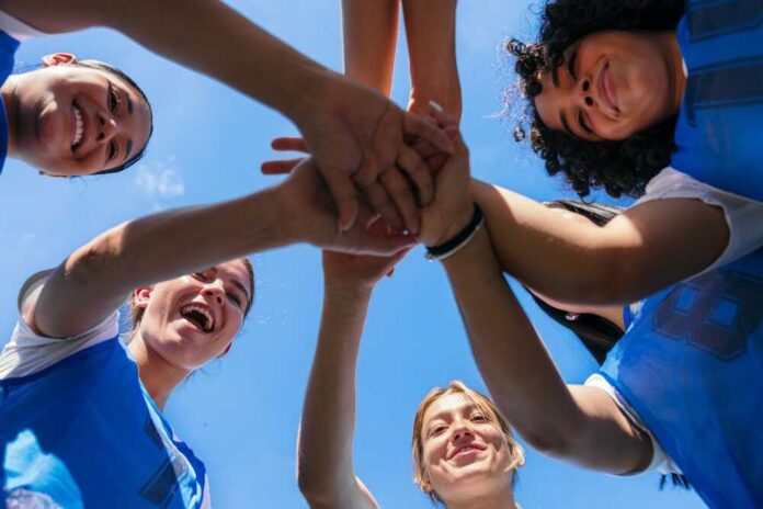 Female,Athletes,Huddling,,Stacking,Hands,,And,Celebrating,Teamwork,Against,A Group of female athletes joining hands in a circle under a blue sky