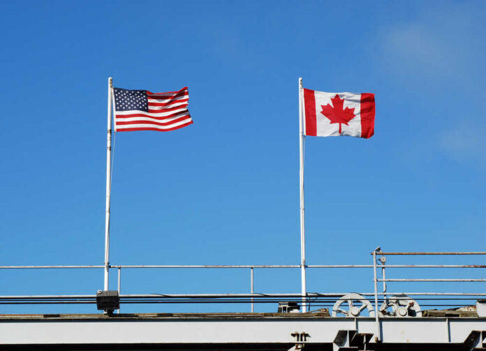 Flags,Flying,At,A,Us,And,Canadian,Border American and Canadian flags flying on poles against a clear blue sky