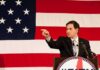 Rubio Torches The “Cheap Import” Myth Speaker in suit pointing during speech before large American flag