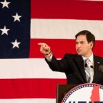 Rubio Torches The “Cheap Import” Myth Speaker in suit pointing during speech before large American flag