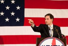 Speaker in suit pointing during speech before large American flag