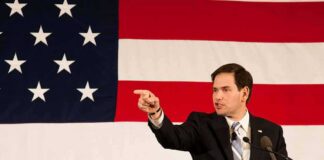 Speaker in suit pointing during speech before large American flag