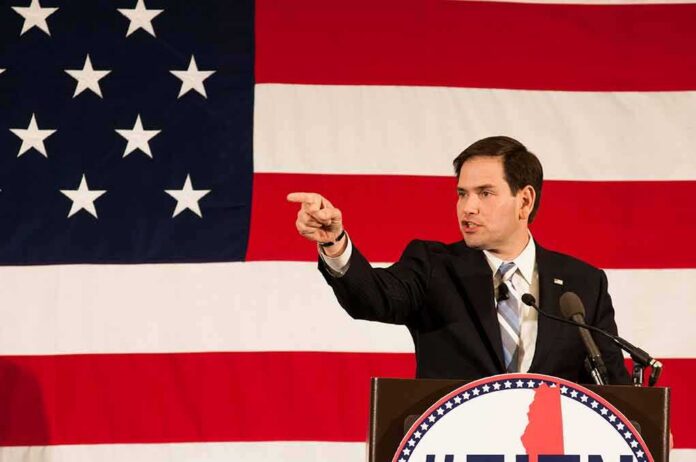 shutterstock_273362126 Speaker in suit pointing during speech before large American flag