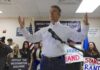 man in white shirt speaking at a rally with supporters and signs behind him