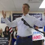 man in white shirt speaking at a rally with supporters and signs behind him