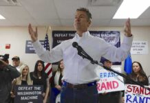 man in white shirt speaking at a rally with supporters and signs behind him