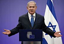man in suit speaking at a podium with Israeli flag