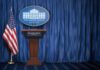 Jobs Surge—Trump Demands Rate Cuts White House press briefing podium with U.S. flag and blue curtain
