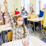 students wearing masks raise hands in a classroom