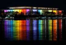 Executive Privilege Shields Kennedy Center Moves Modern building lit in rainbow colors reflected on water at night