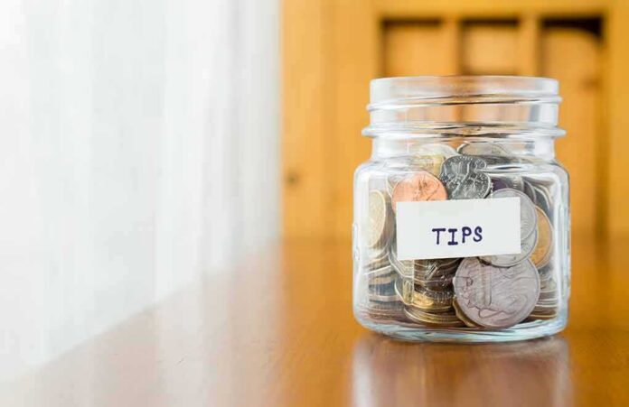 shutterstock_223702975 Tip jar filled with assorted coins on a wooden counter