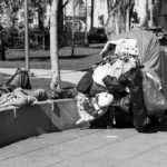 Progressive Paradise Admits Failure – Now What? Man sleeping on park ledge beside overloaded cart